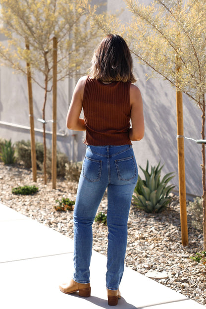 Model wears the Free People Brown Tilly Vest Top on a sidewalk, paired with blue jeans, facing away by plants.