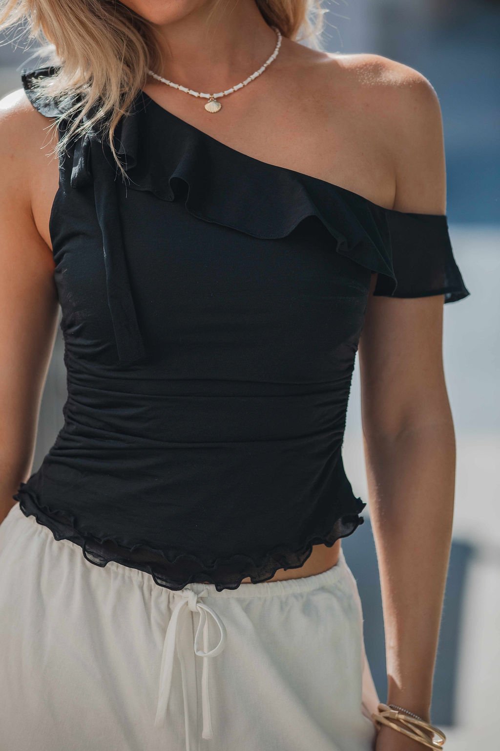 Woman in a black Free People Isla Off The Shoulder Top - Black-FINAL SALE, styled with white pants and chic accessories.