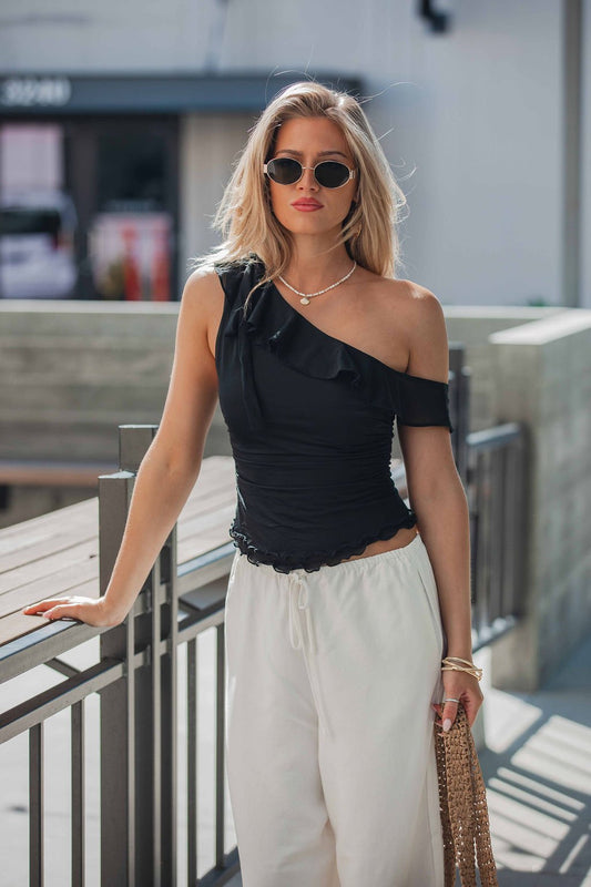 Woman in sunglasses wears a black Free People Isla Off The Shoulder Top, white pants, and carries a woven bag.