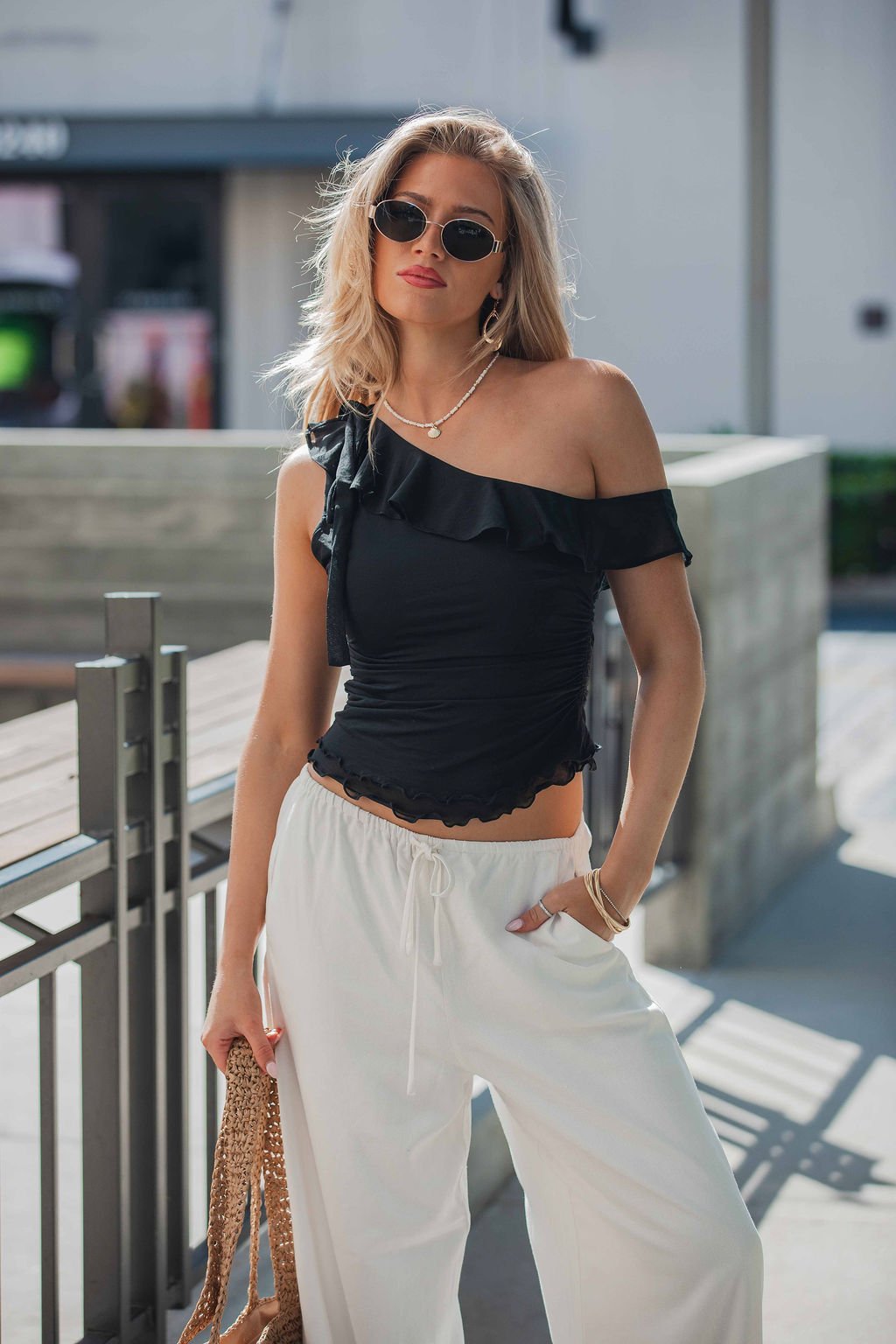 Woman in sunglasses and white pants poses confidently in a Free People Isla Off The Shoulder Top - Black, holding a woven bag.
