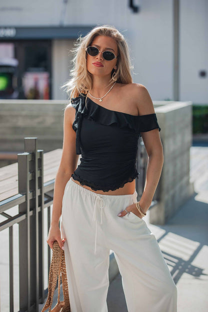 Woman in sunglasses and white pants poses confidently in a Free People Isla Off The Shoulder Top - Black, holding a woven bag.