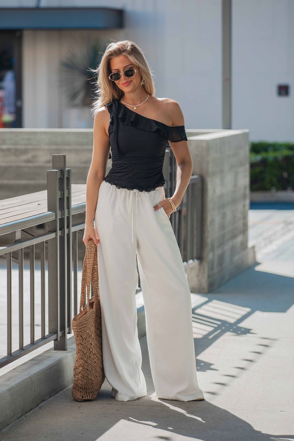 Woman wearing a black Free People Isla Off The Shoulder Top, white wide-leg pants, sunglasses, and a woven bag while outdoors.