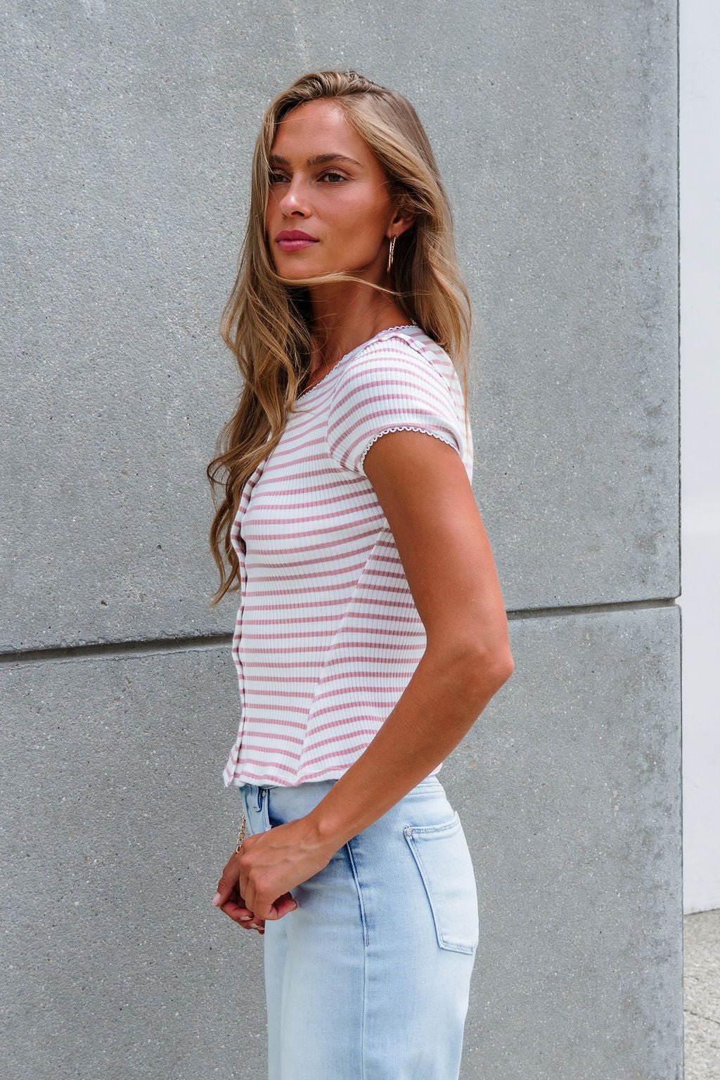 Woman with long hair in the Free People Stripe Most Wanted Top - Mauve stands by a gray concrete wall in light jeans.