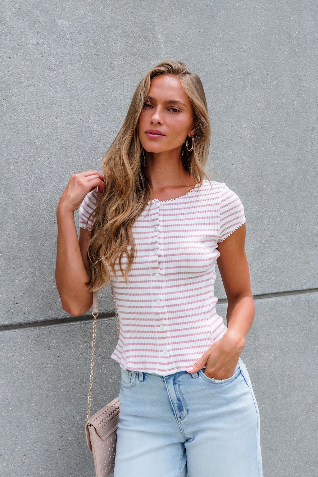 Woman with long blonde hair wears the Free People Stripe Most Wanted Top - Mauve, styled with light jeans against a gray concrete wall.