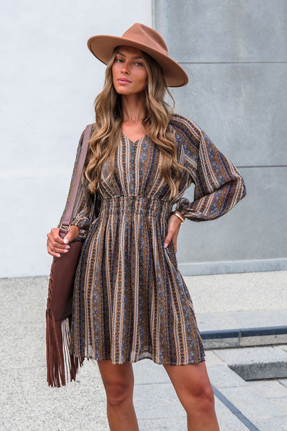 Woman in Genevieve Floral Striped Mini Dress—Mocha, with balloon sleeves, wide-brim hat, brown fringe bag, wavy hair, neutral expression.