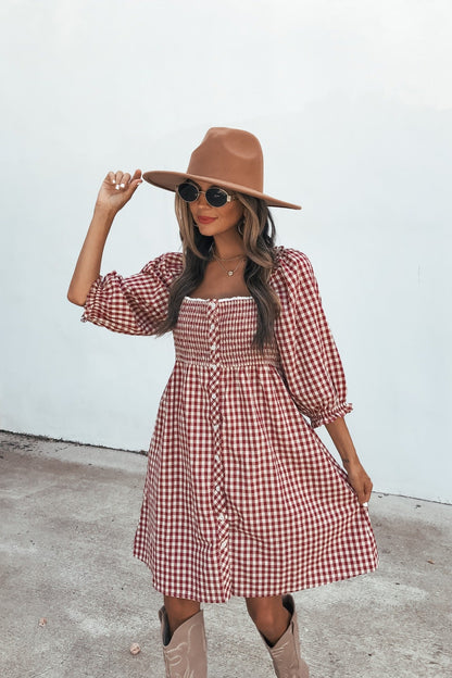 Woman in a Gingham Babydoll Mini Dress - Burgundy, tan hat, and sunglasses poses outdoors on concrete, holding her hat.
