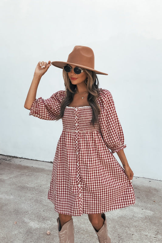 Woman in a Gingham Babydoll Mini Dress - Burgundy, tan hat, and sunglasses poses outdoors on concrete, holding her hat.