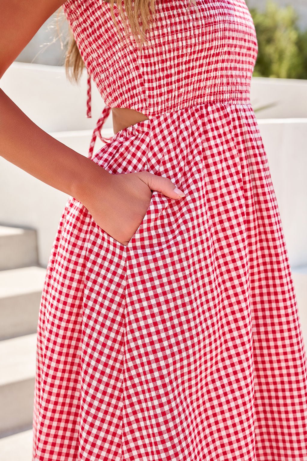 Woman wearing the Gingham Open Back Midi Dress in red, standing outdoors with her hand in a pocket near steps.