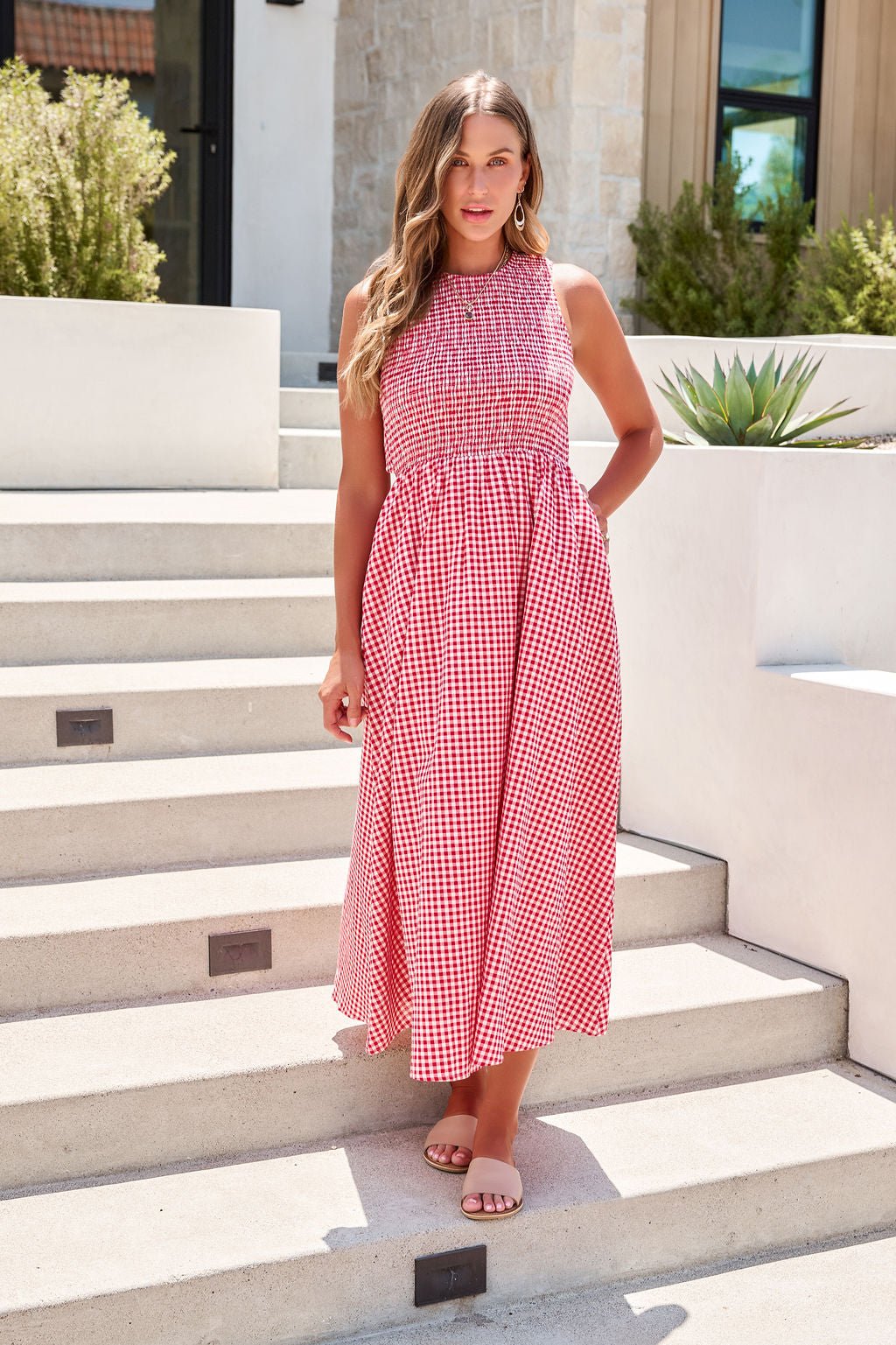 Woman wears the Gingham Open Back Midi Dress - Red, standing outdoors on steps with plants and a modern building behind her.