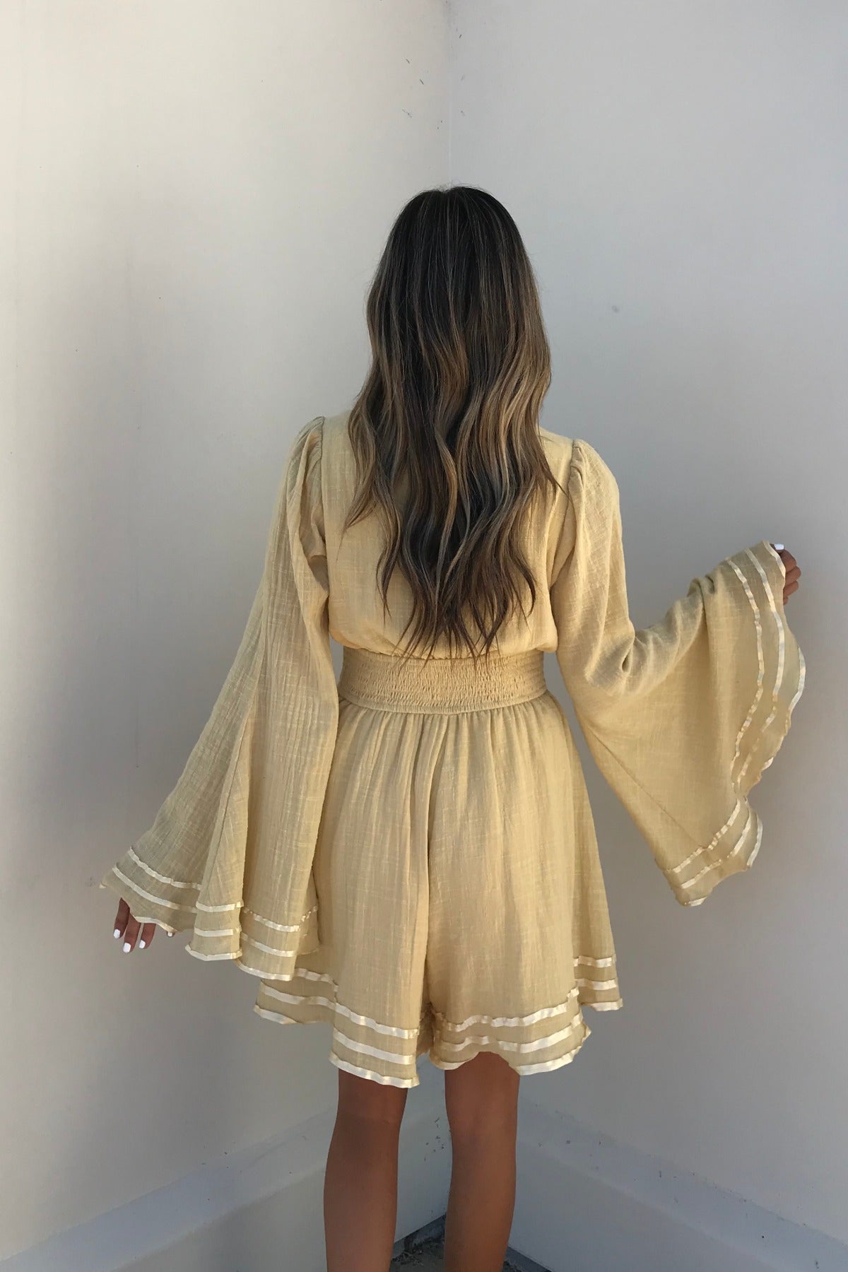 A woman with wavy hair wears the light yellow Golden Hour Bell Sleeve Romper - FINAL SALE, featuring flared sleeves and shorts.
