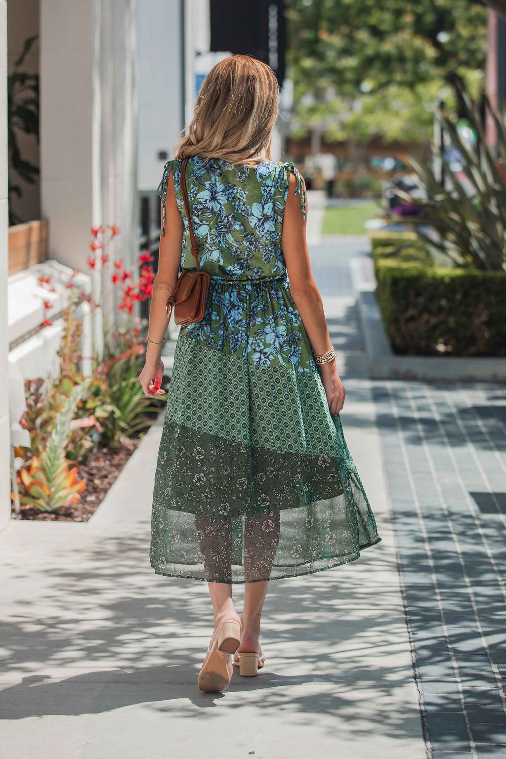 Woman in the Greenhaven Print Midi Dress walks on a sunny sidewalk, her long hair flowing and sandals completing her look.