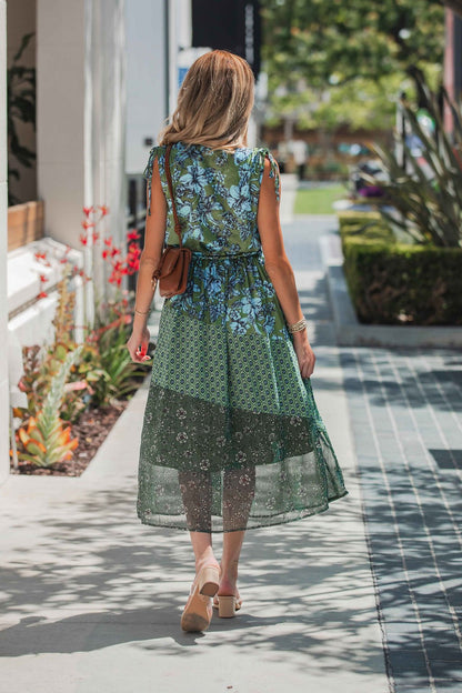 Woman in the Greenhaven Print Midi Dress walks on a sunny sidewalk, her long hair flowing and sandals completing her look.