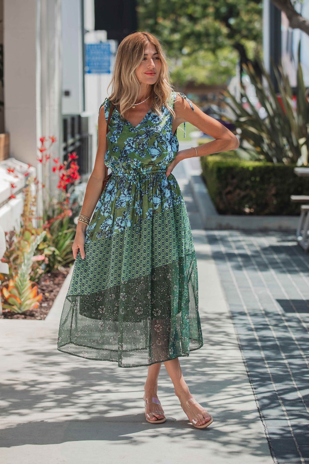 Woman wears the Greenhaven Print Midi Dress - FINAL SALE on a sunny sidewalk with plants, styled with sandals and bracelets.