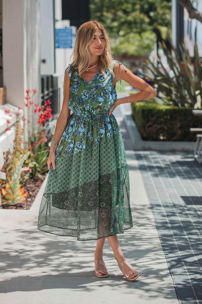 Woman wears the Greenhaven Print Midi Dress - FINAL SALE on a sunny sidewalk with plants, styled with sandals and bracelets.