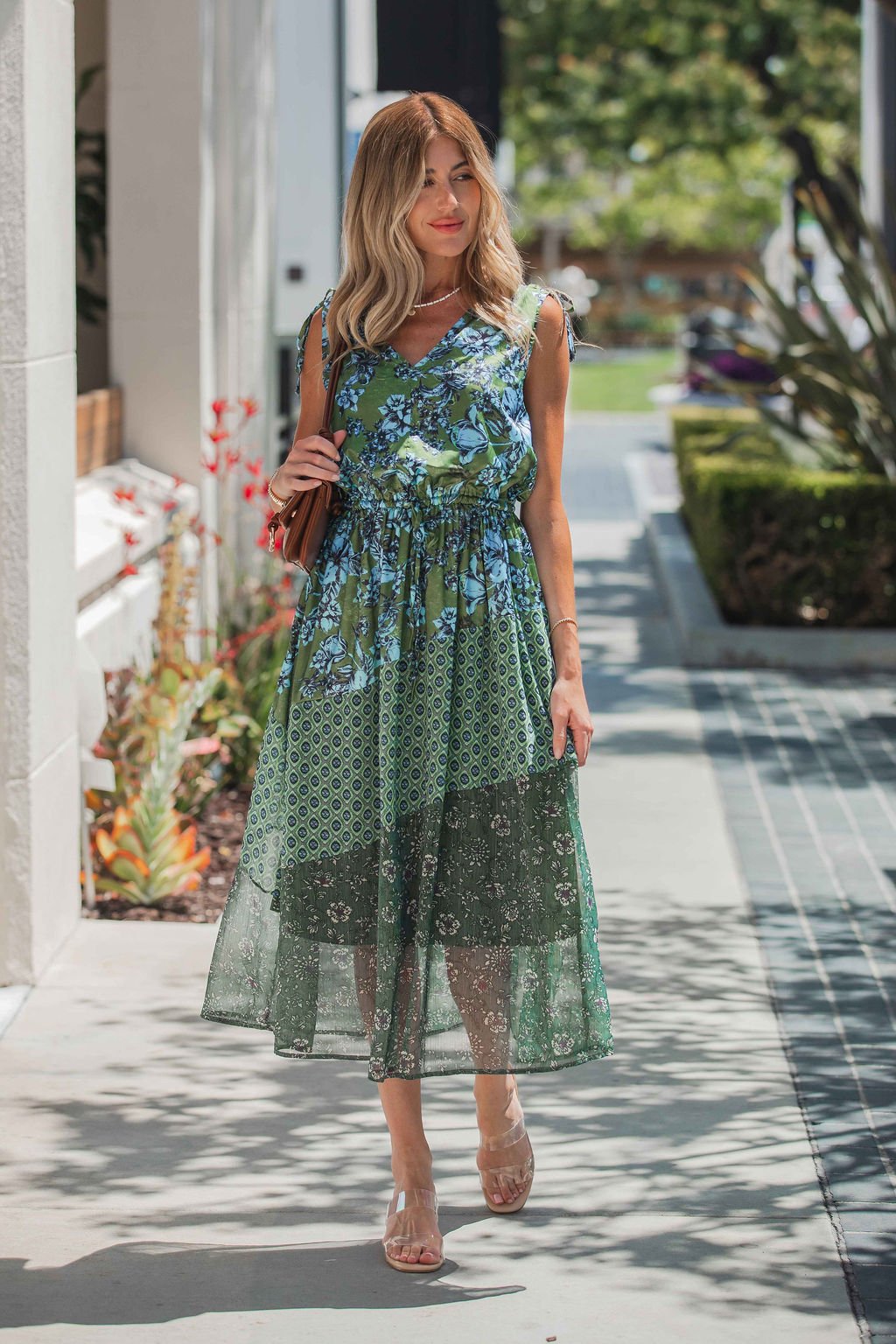 Woman in a Greenhaven Print Midi Dress - FINAL SALE walks outdoors on a sunny day, carrying a brown purse and wearing clear-strap sandals.