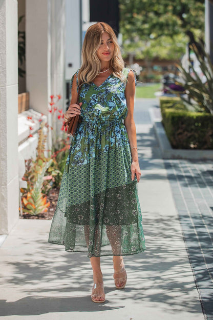 Woman in a Greenhaven Print Midi Dress - FINAL SALE walks outdoors on a sunny day, carrying a brown purse and wearing clear-strap sandals.