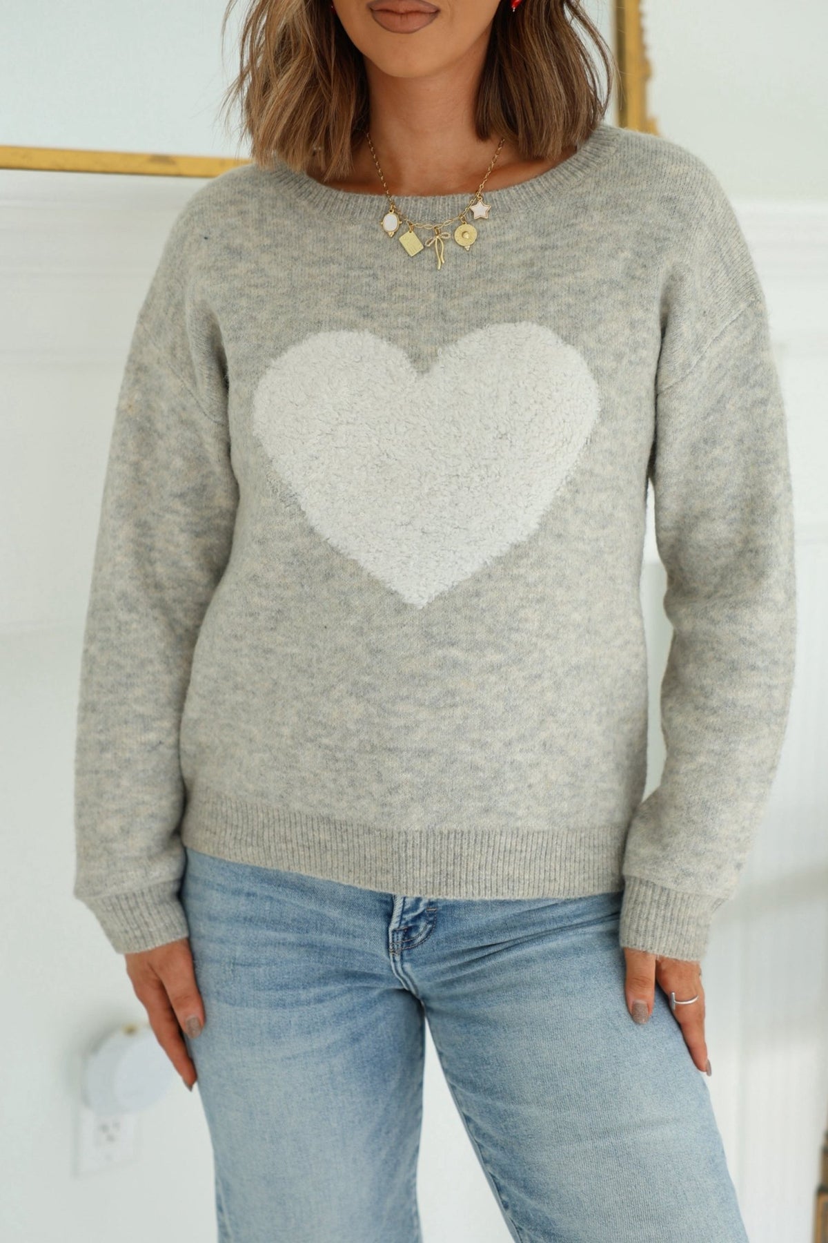 Woman wearing a Grey and White Heart Shape Sweater, blue jeans, gold necklace, and rings.