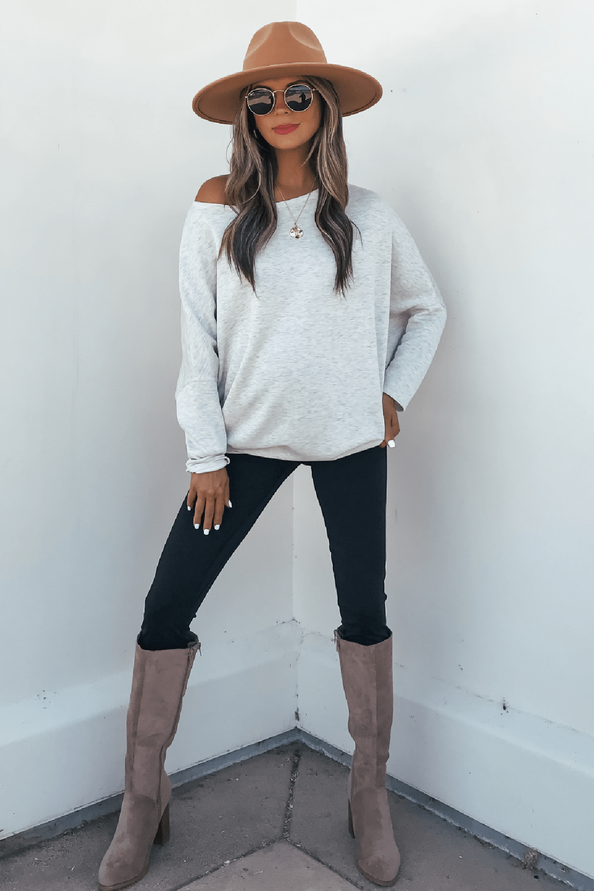 Woman in a hat, sunglasses, Grey Soft Cloud Scuba Pullover, black pants, and taupe boots stands against a white background.