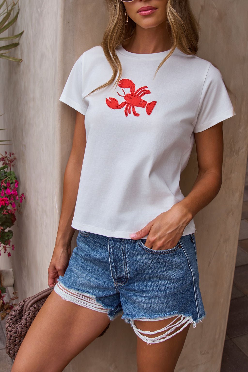 Woman in a Harbor Lobster Patch Tee - FINAL SALE and ripped denim shorts, exuding a cool coastal vibe against the wall.
