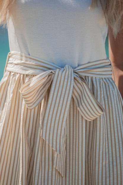 Woman in the Harbor Stripe Tie Midi Dress - Taupe, featuring a large waist bow, stands outdoors in a breezy coastal-inspired outfit.