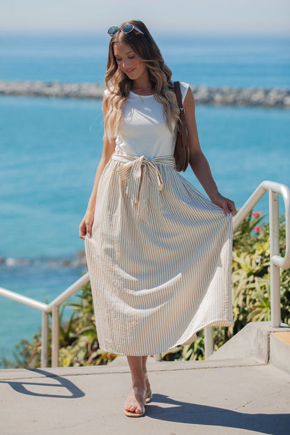 A woman in the Harbor Stripe Tie Midi Dress - Taupe stands by the ocean, holding her skirt on a sunny day.