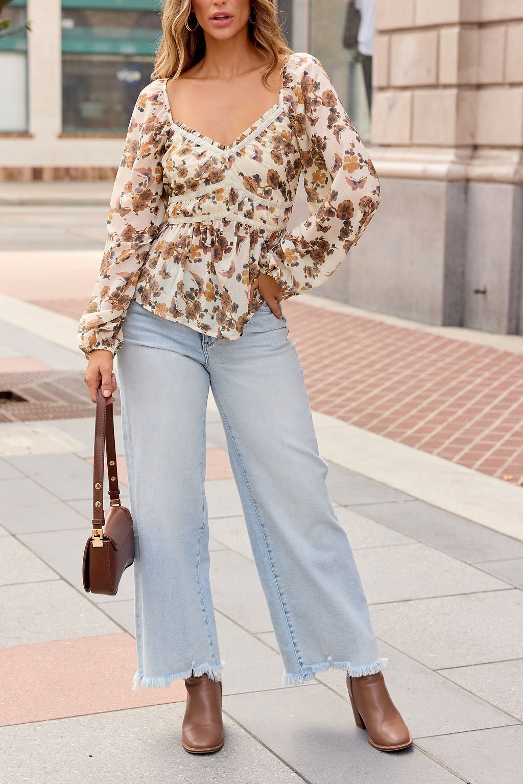 Woman in Hazel Floral Print Top - Cream, light blue wide-leg jeans, brown boots, and a brown handbag on a city sidewalk.