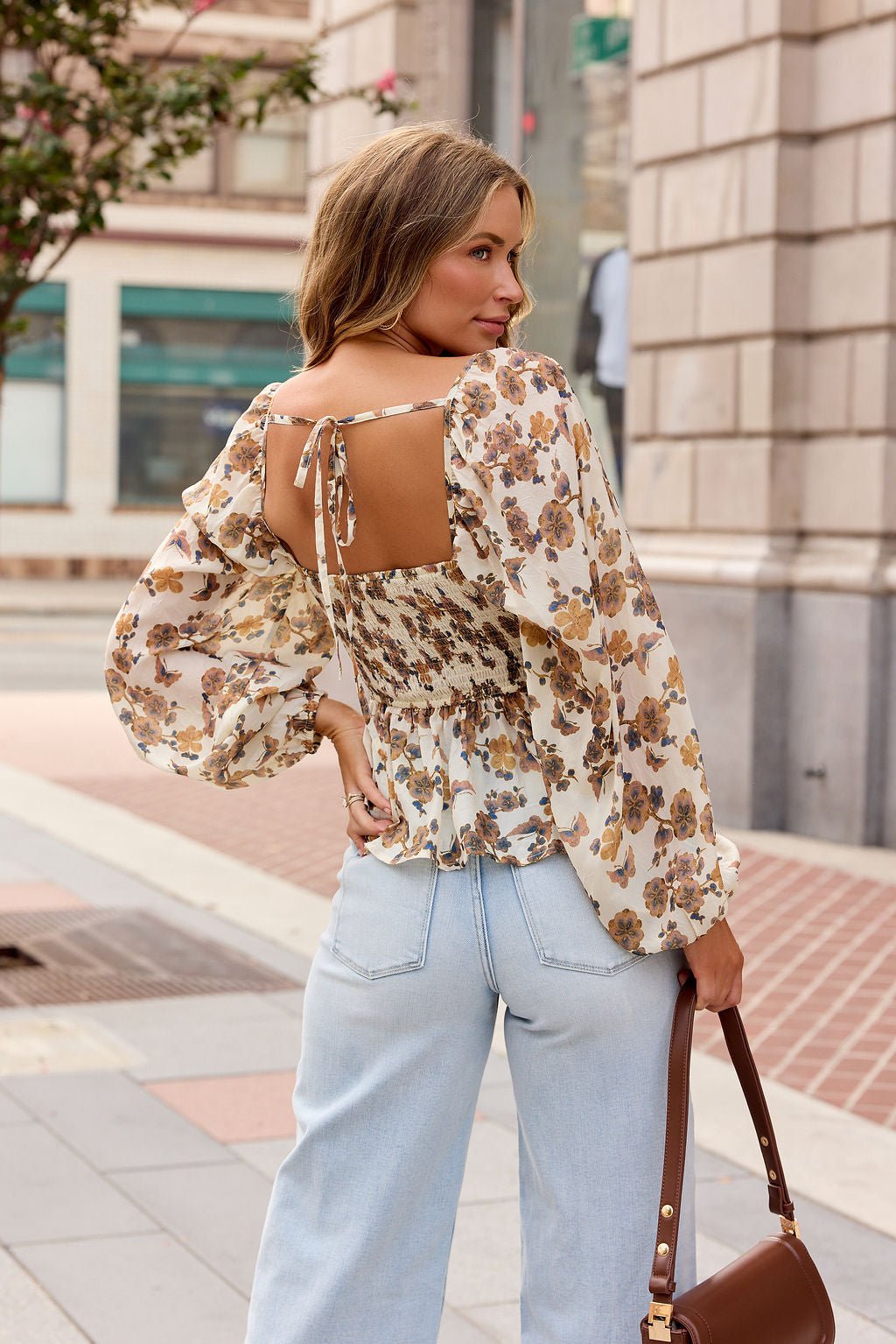 Woman in a Hazel Floral Print Top - Cream, light jeans, and brown handbag stands outdoors, facing away on a city sidewalk.