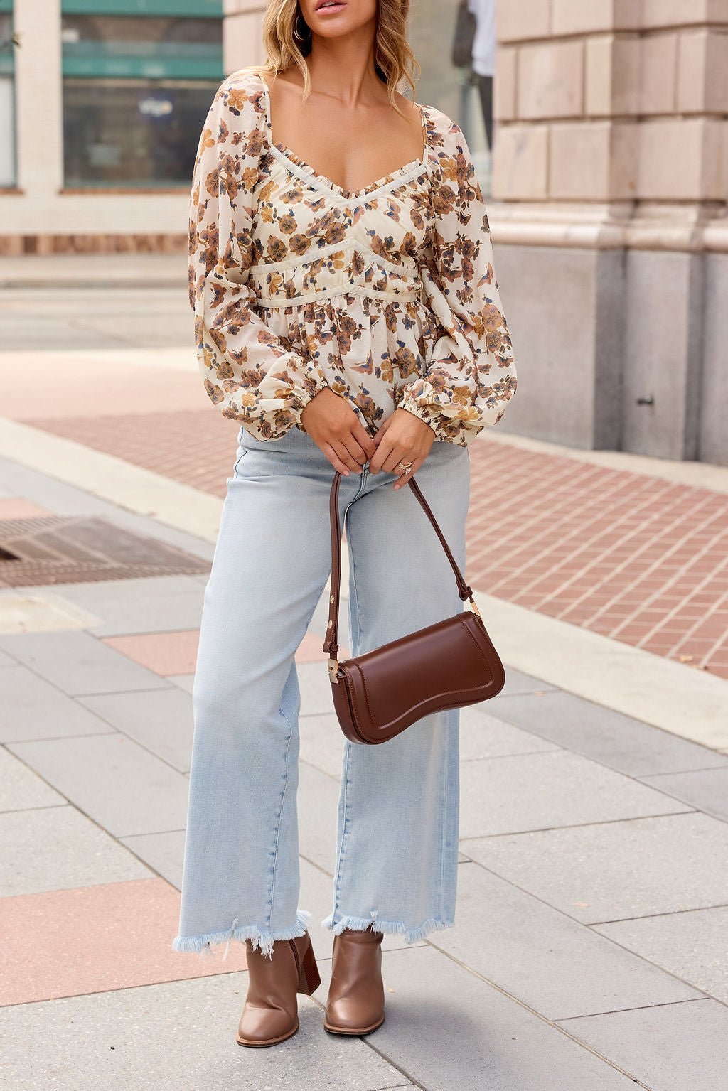 Woman in a Hazel Floral Print Top - Cream, light jeans, brown boots, and handbag stands on a city sidewalk.