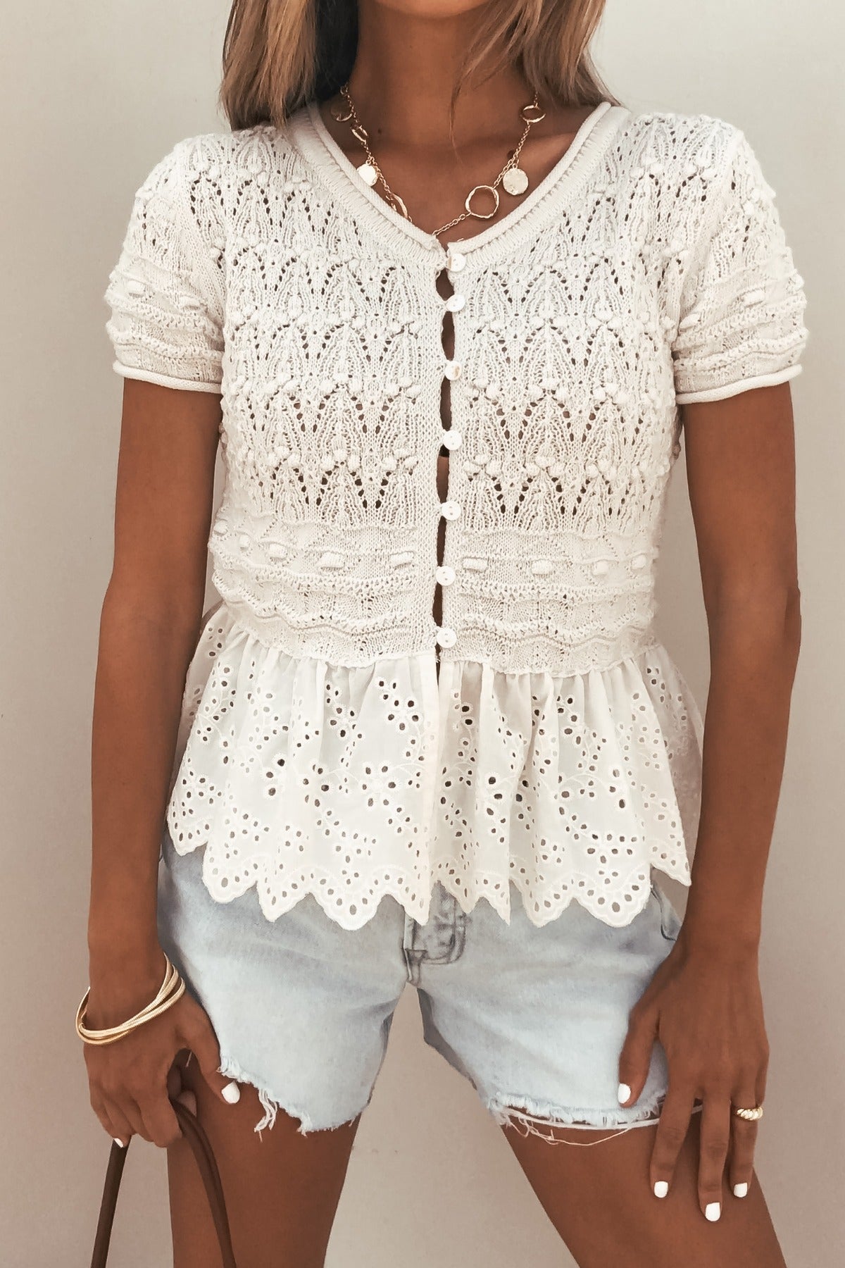 Woman wears the Ivory Crochet Peplum Top with light denim shorts and gold jewelry.