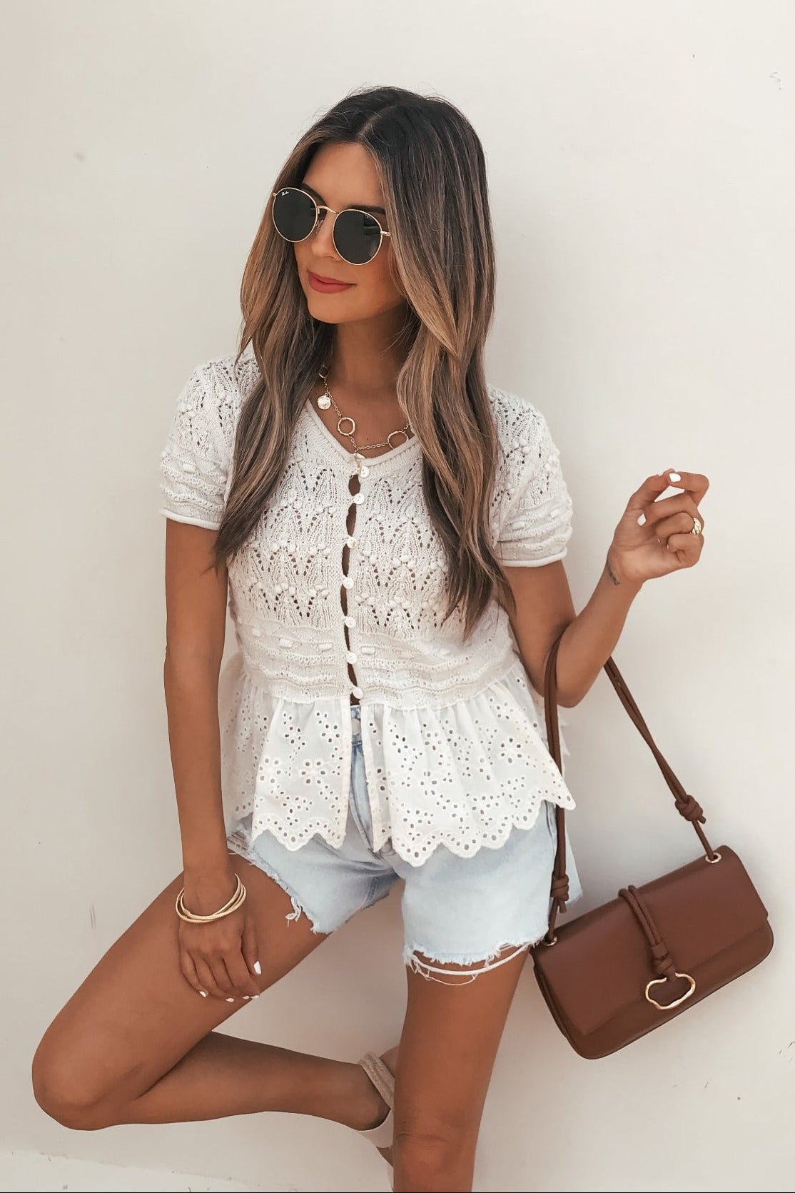 Woman in sunglasses wears an Ivory Crochet Peplum Top, denim shorts, and holds a brown handbag against a plain background.