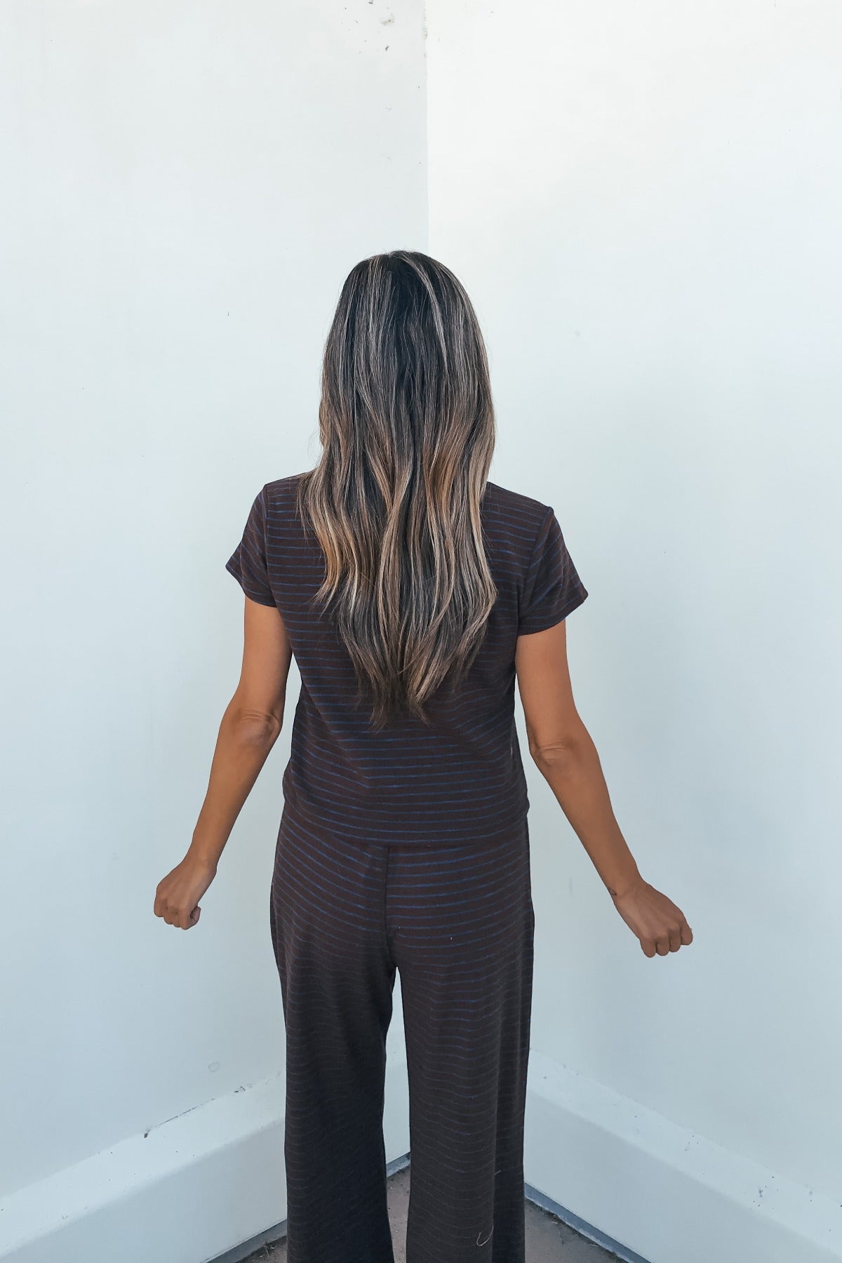 Woman with long wavy hair wears the Kacey Striped Short Sleeve Top - Brown, standing facing a white wall with arms at her sides.