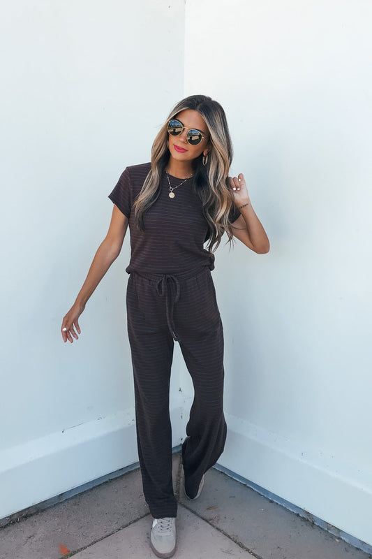 Woman in sunglasses wears the Kacey Striped Short Sleeve Top in brown and sneakers, smiling with one hand raised against a white wall.