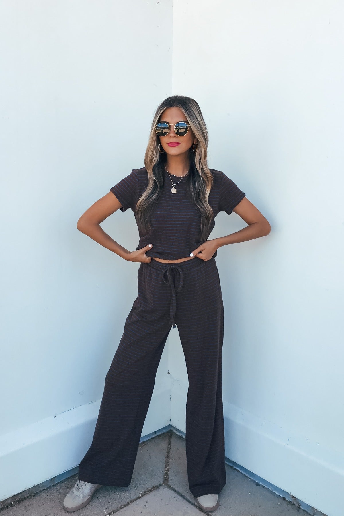 Woman in sunglasses and Kacey Striped Short Sleeve Top - Brown poses confidently against a white wall, hands on hips.