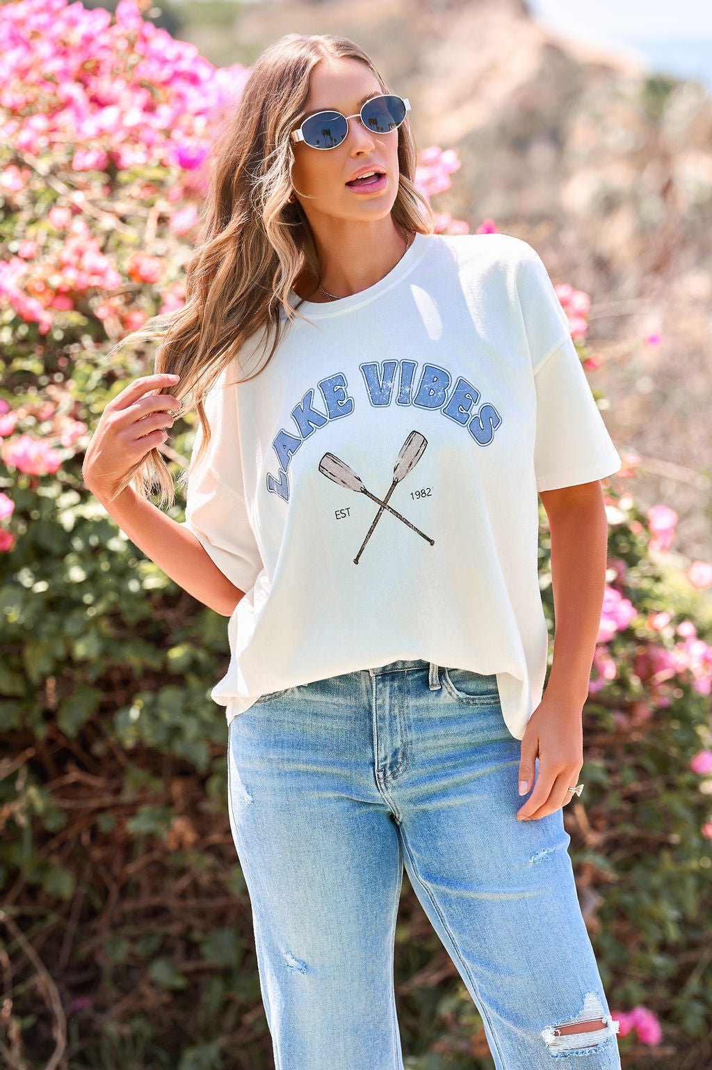 Woman in sunglasses and an Ivory Lake Vibes Graphic Tee stands by pink flowers, wearing light blue jeans on a sunny day outdoors.