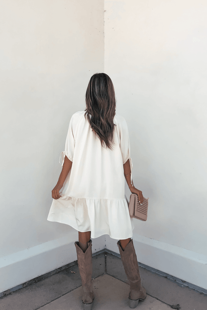 Woman in a Laurie Cream Button Down Mini Dress-FINAL SALE and brown boots holds a beige studded clutch, facing the corner.
