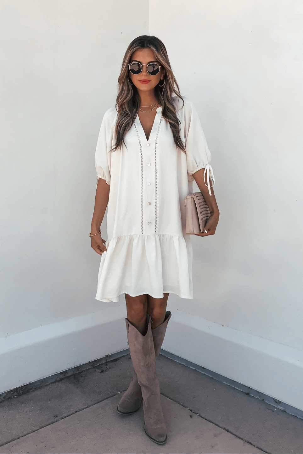 Woman in the Laurie Cream Button Down Mini Dress stands by a white wall, styled with boots, sunglasses, and a beige clutch.