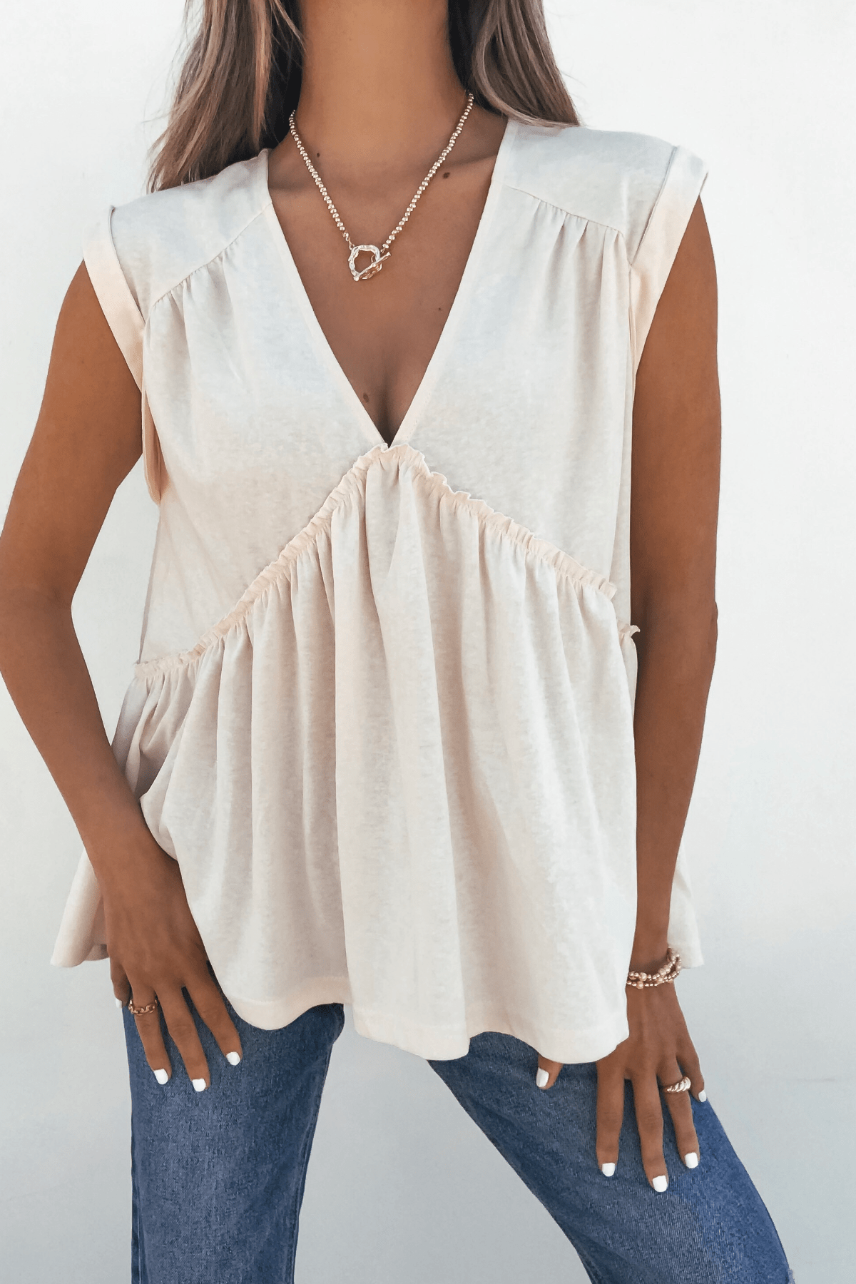 Model wears Lena’s Carefree Afternoon Cream Babydoll Top with ruffles, blue jeans, and gold jewelry against a white background.