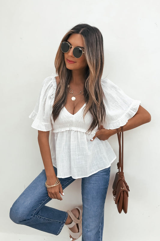 Woman in sunglasses wears the Lola White Ruffled Babydoll Top with blue jeans and sandals, posing against a white wall.