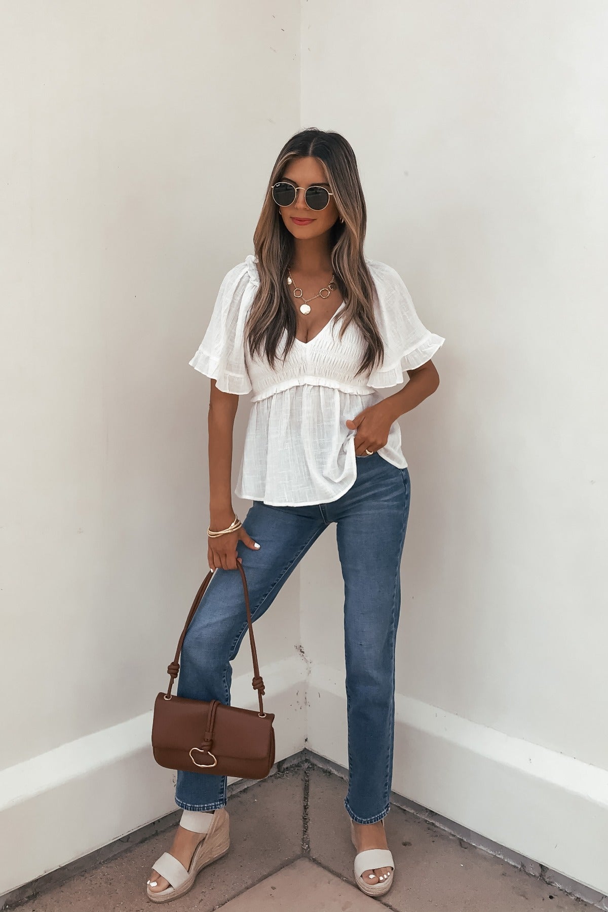 Woman in sunglasses wears the Lola White Ruffled Babydoll Top, blue jeans, beige heels, and holds a brown handbag.