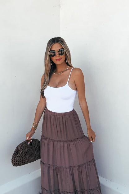 Woman in sunglasses wears a white tank and the Lori Ruffle Tiered Maxi Skirt - Mocha, holding a woven bag by a white wall.
