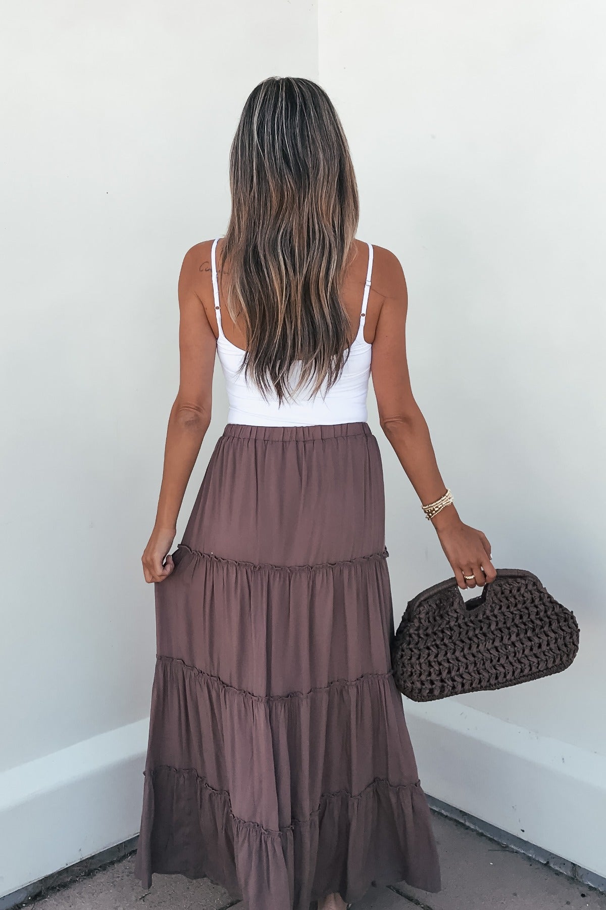 Woman in Lori Ruffle Tiered Maxi Skirt - Mocha holds a woven bag, facing away against a white wall.