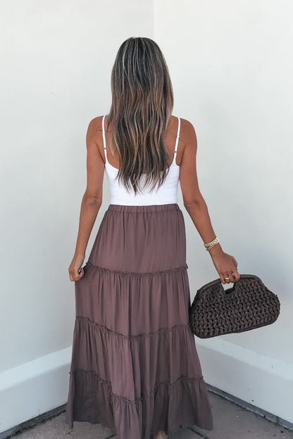 Woman in Lori Ruffle Tiered Maxi Skirt - Mocha holds a woven bag, facing away against a white wall.