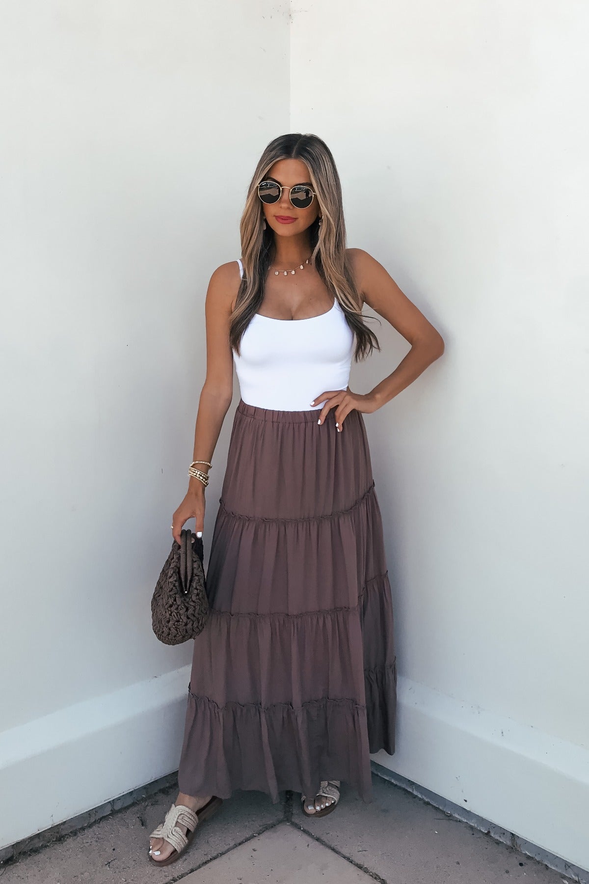 Woman in sunglasses, white tank, and Lori Ruffle Tiered Maxi Skirt - Mocha, holding a textured bag by a white wall.