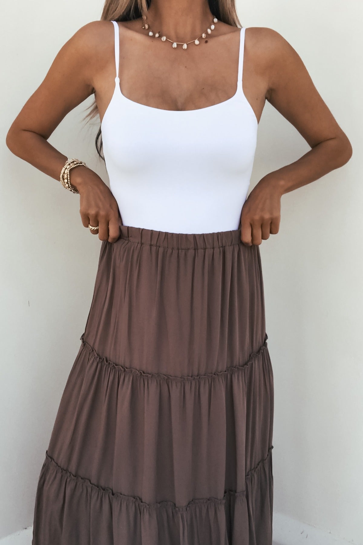 Woman in a white tank top and the Lori Ruffle Tiered Maxi Skirt - Mocha, styled with gold bracelets and a beaded necklace.