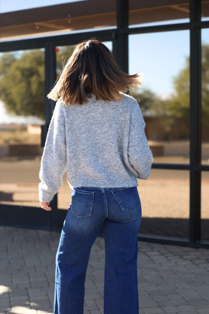 Woman with shoulder-length hair in the Lucille Grey Button Down Sweater Cardigan and blue jeans stands outdoors, facing away.