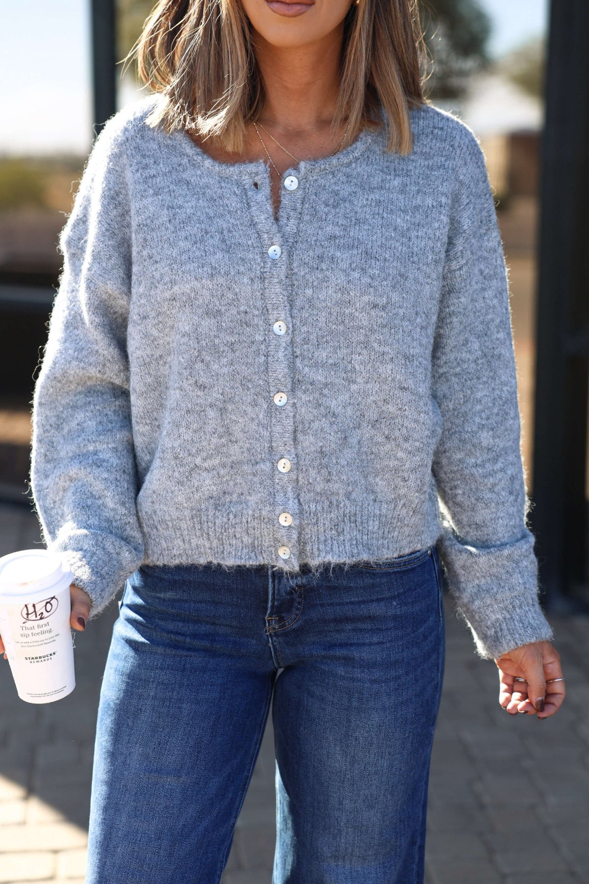 Woman in a Lucille Grey Button Down Sweater Cardigan and blue jeans holds a white coffee cup outdoors.