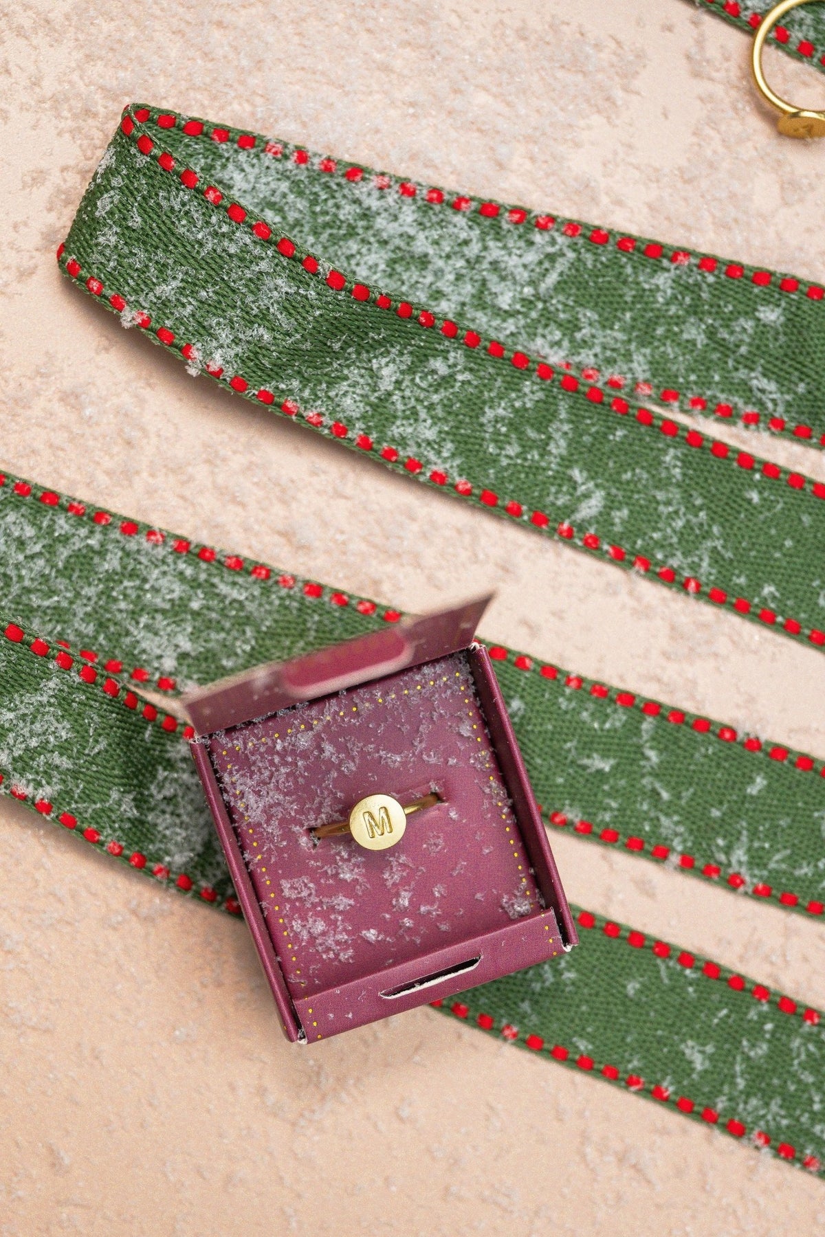 The Lucky Feather Gold Initial Pendant Ring - FINAL SALE sits in an open box by a green ribbon with red edges on snow.