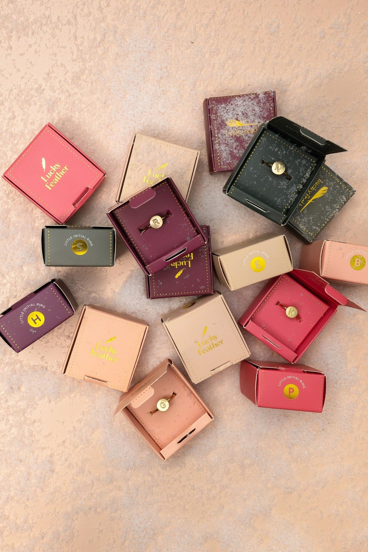 Lucky Feather Gold Initial Pendant Ring on pink textured surface with colorful jewelry boxes and gold initial pins.