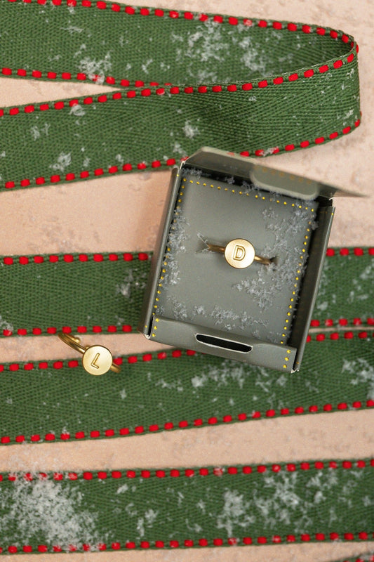 Two Lucky Feather Gold Initial Pendant Rings with “L” and “D” engravings rest on faux snow with green ribbon. Perfect personalized gift.