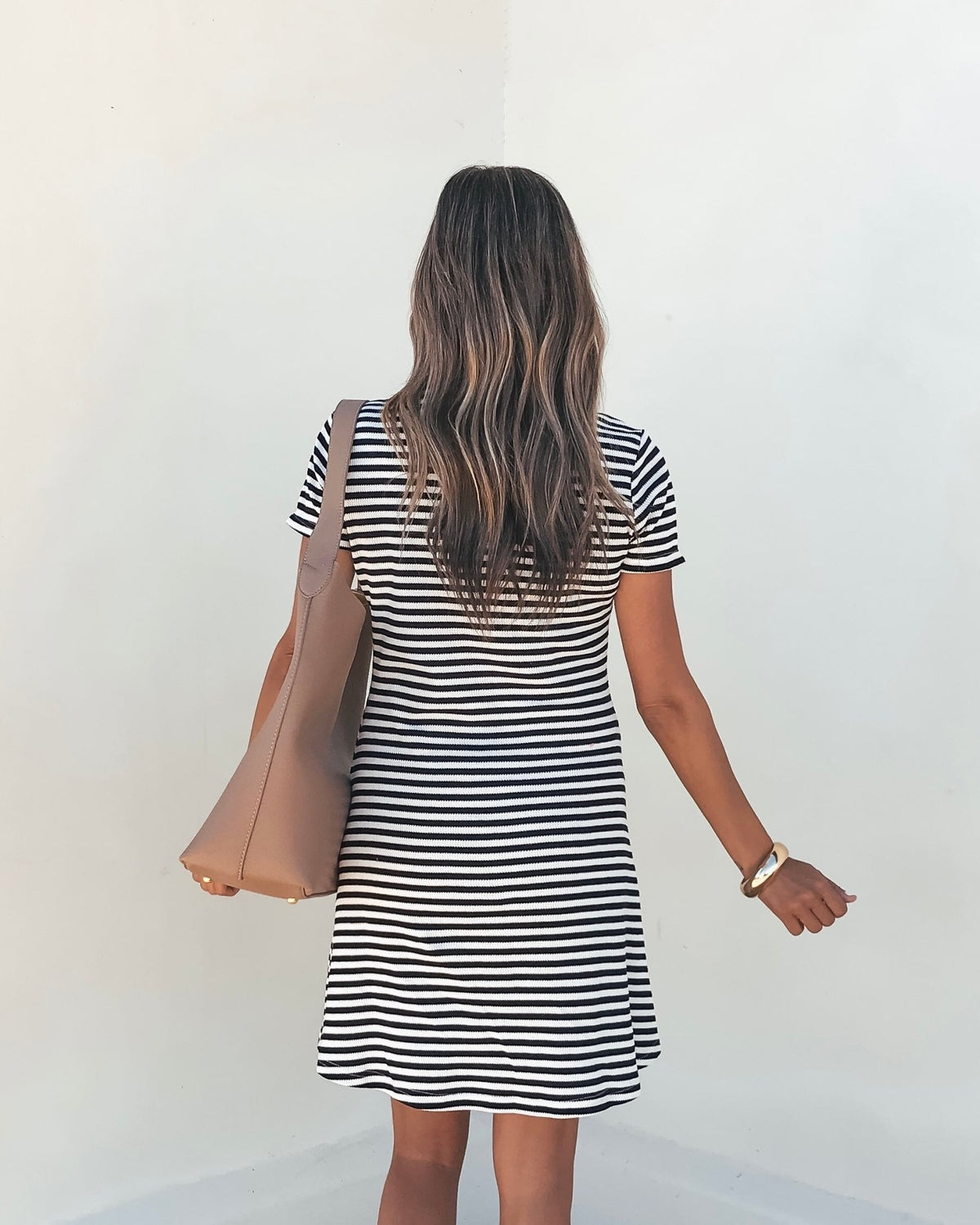 Woman with long wavy hair wears the Lucy Black and White Striped Mini Dress-FINAL SALE, holding a tan bag by a white wall.